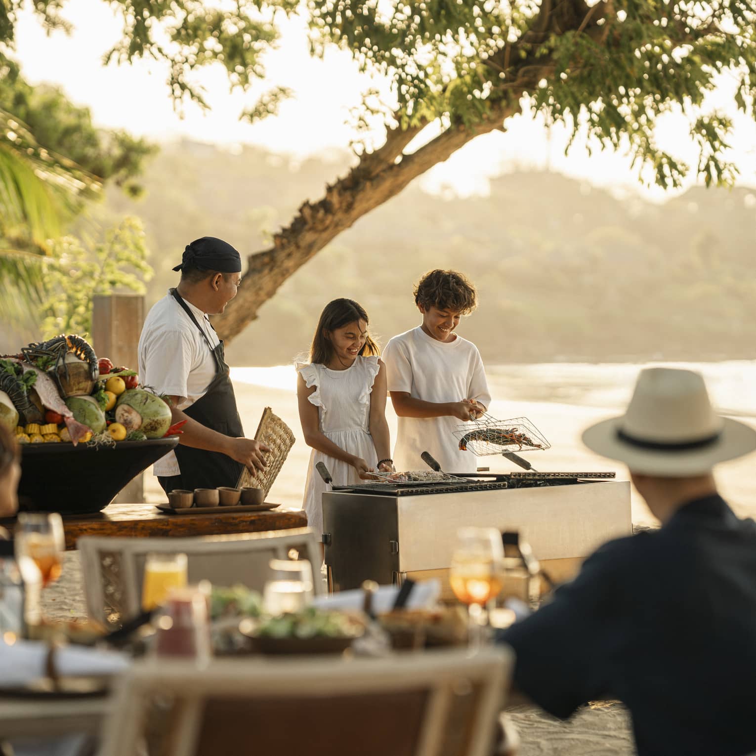A chef smiles at two children who barbecue shellfish in a grilling basket on the beach as their parents watch.