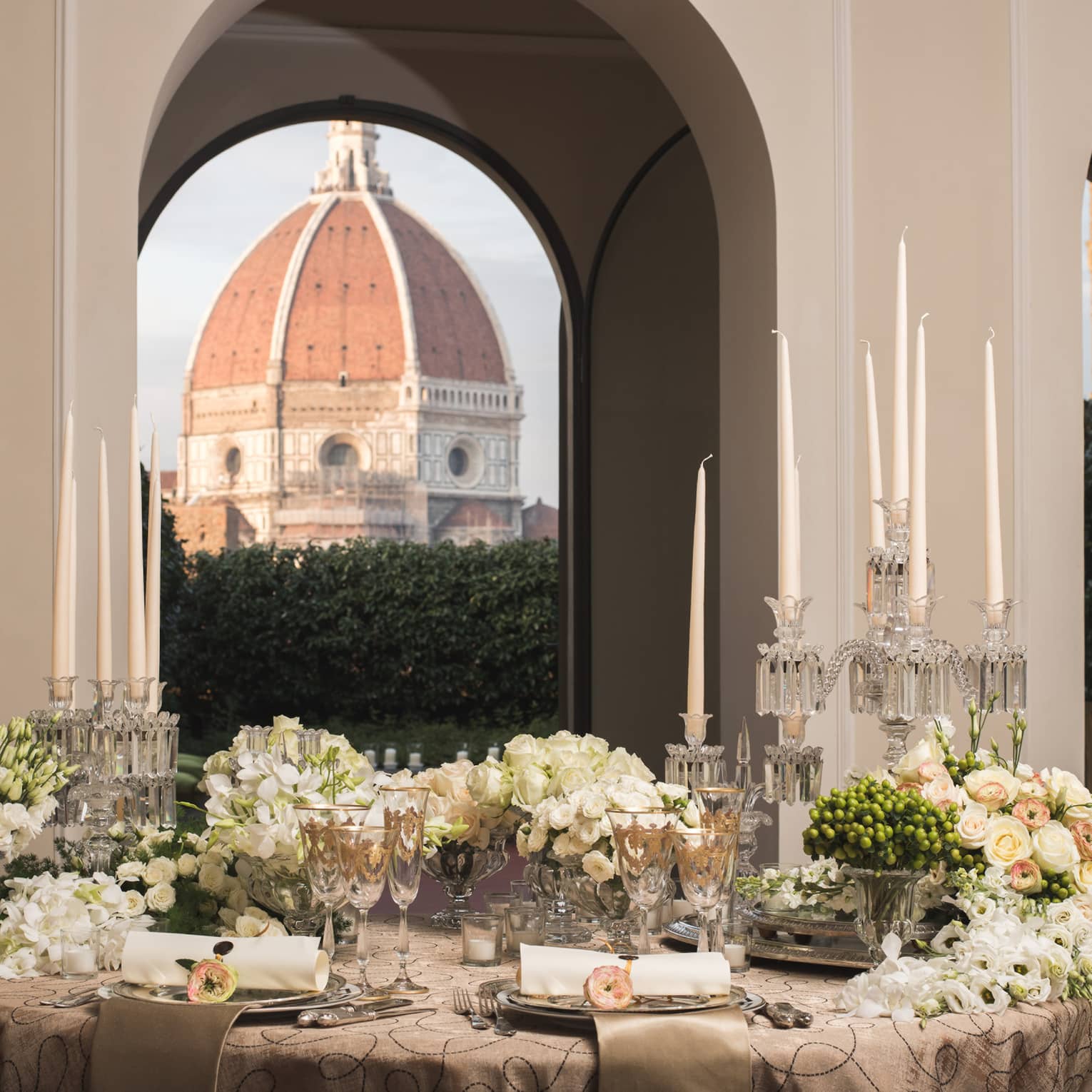 Elegant dining table set with candles, flowers, and glassware, with a large dome visible through arched windows in the background.