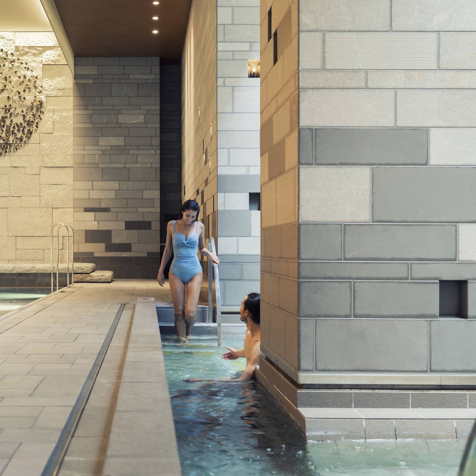 A guest enters a pool via the stairs to meet another guest who is half-hidden by a large grey-tiled column in the foreground.