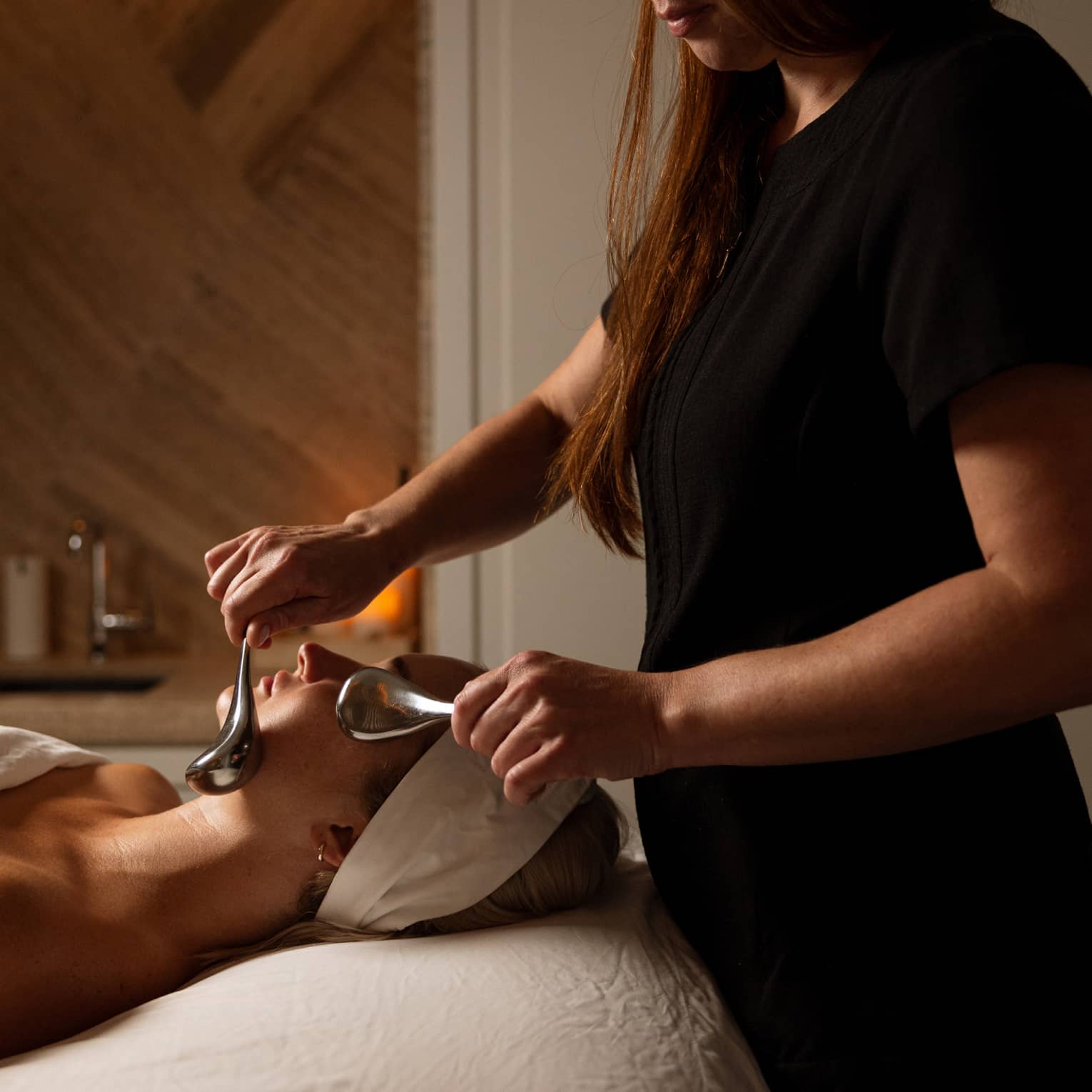 A guest draped in white and wearing a white headband lies on a treatment table in a softly lit spa treatment setting while an aesthetician applies a facial treatment