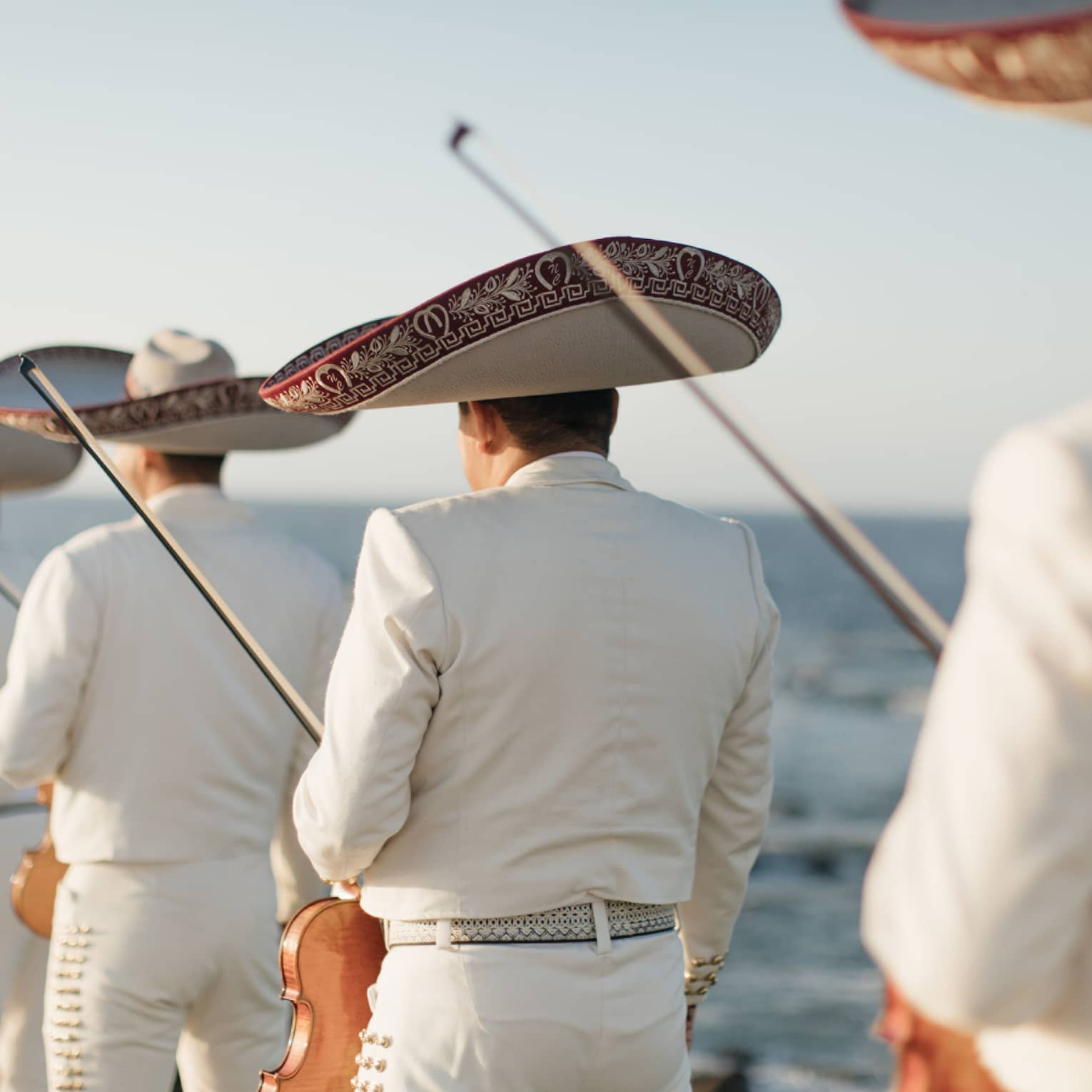 The backs of four men in white suits and large hats holding violins with the ocean in the background.