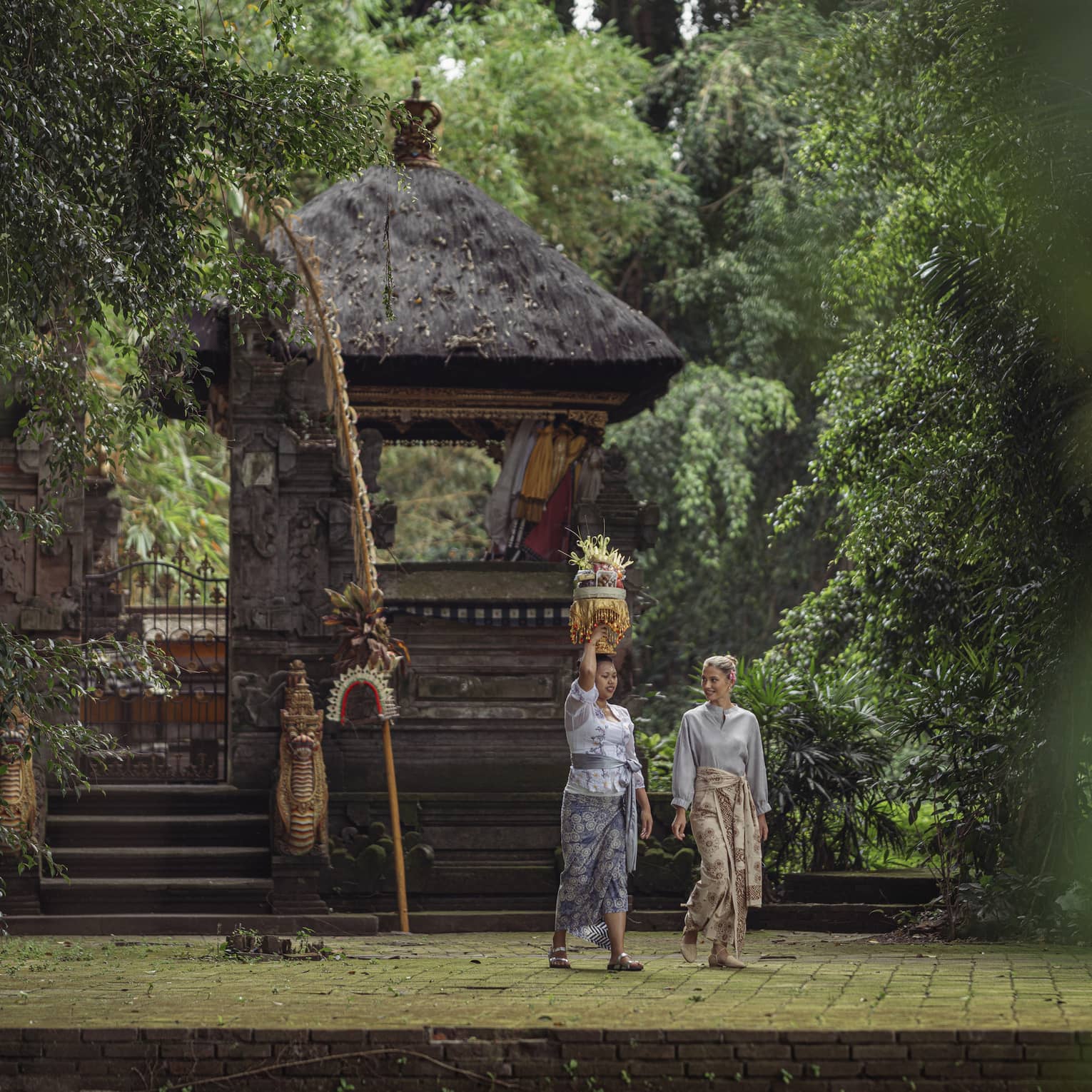 Two women wearing batik skirts walk by a temple in a lush tropical area as one of them carries a golden tray on her head.