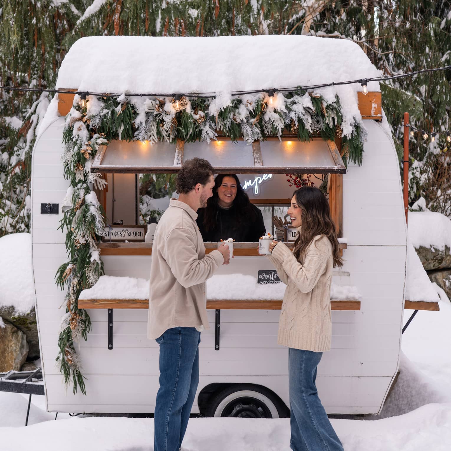 A couple holds mugs of hot chocolate outside of a camper in a snowy courtyard