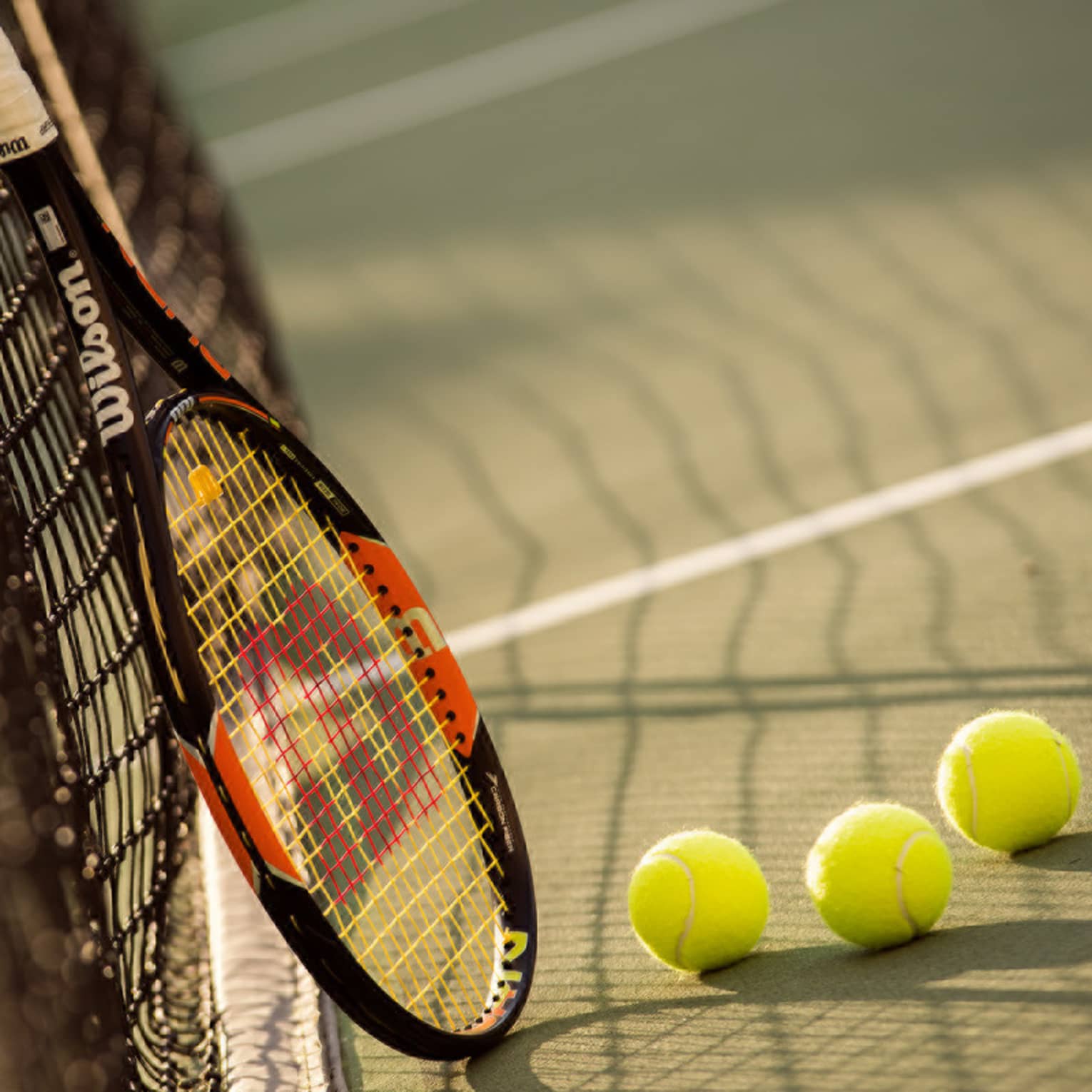 Red and black tennis racket leans against net by green tennis balls on court