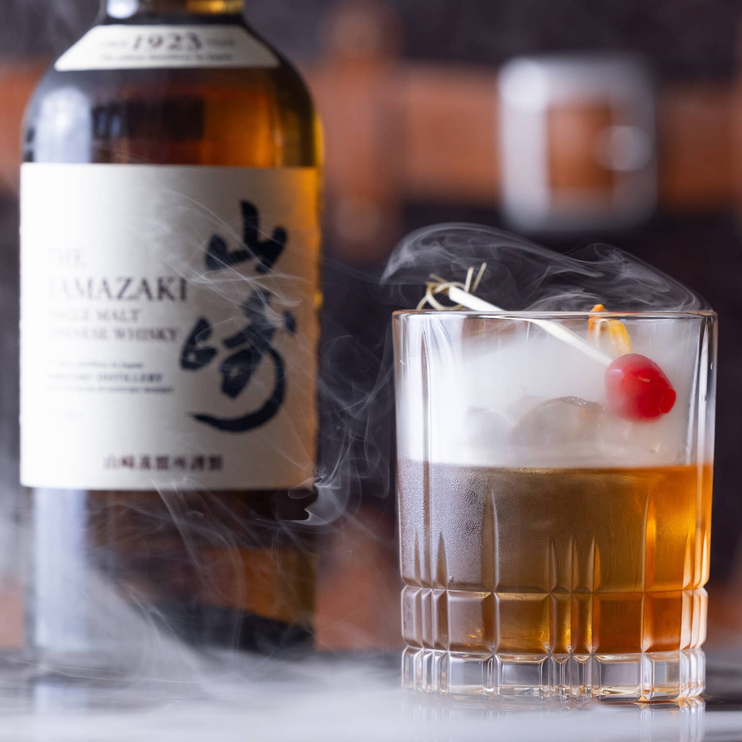 Bottle of Japanese whisky beside half-full cut-crystal glass with red cherry garnish, dry ice mist swirling from the glass.