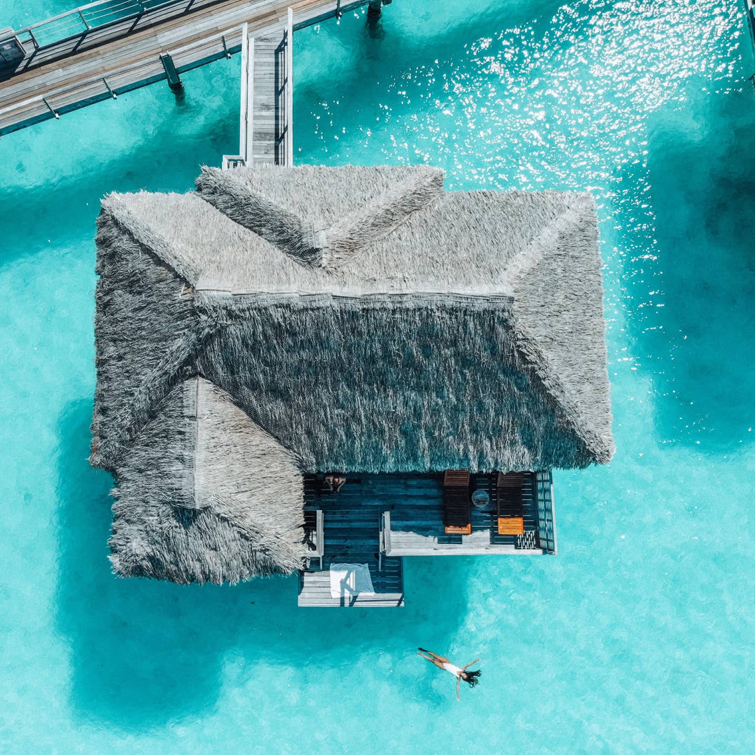 Aerial view of person swimming away from overwater bungalow in turquoise lagoon