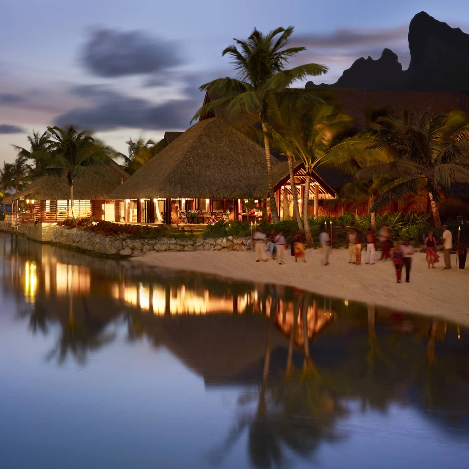 Guests on sand beach by pavilions with lights at night