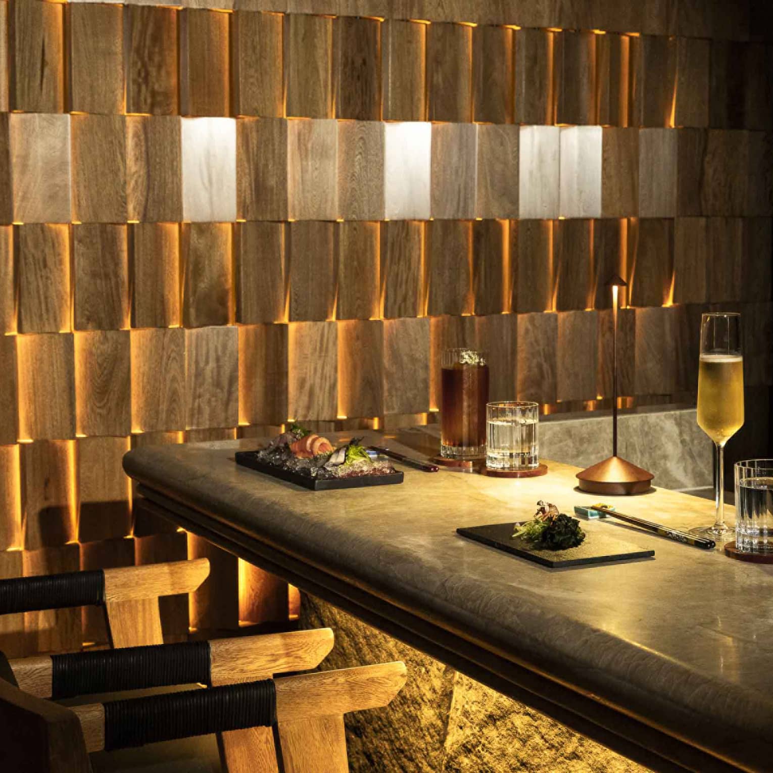 An artistic dining space with soft, warm light, a long table with seating and a wall of backlit wood tiles