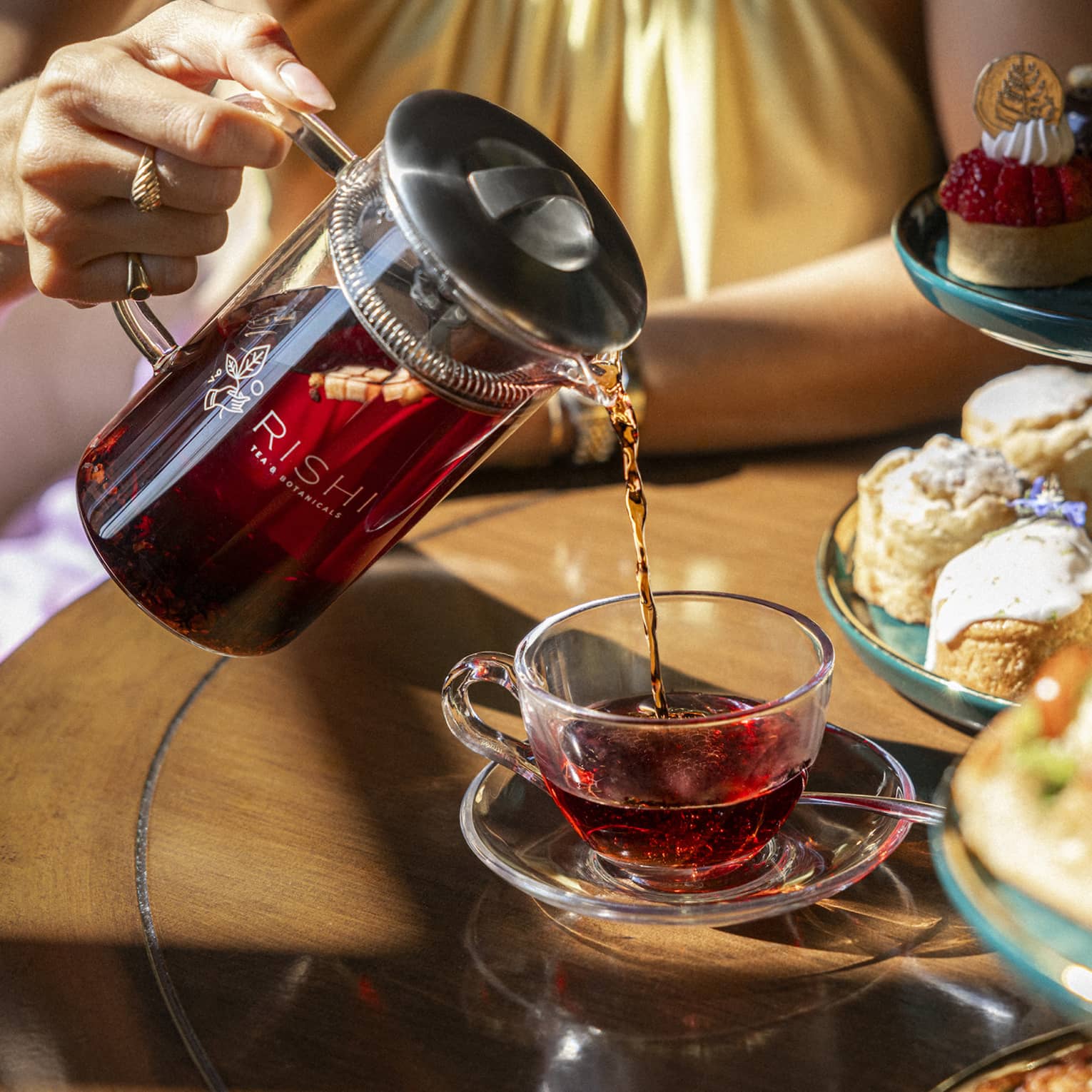 Person pouring tea from a tea kettle into a mug, with various desserts beside it.