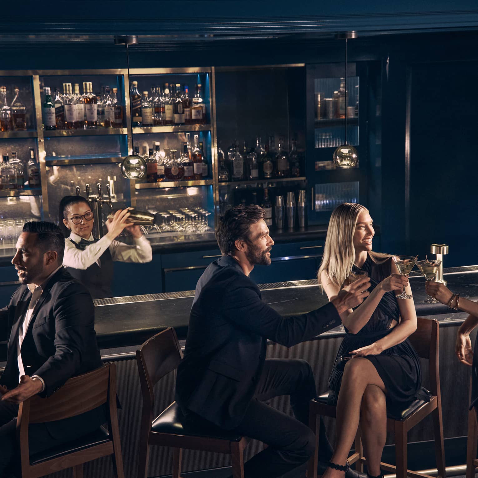 Guests chat over drinks at the bar at Prosperous Penny as a bartender shakes a cocktail shaker behind the bar.