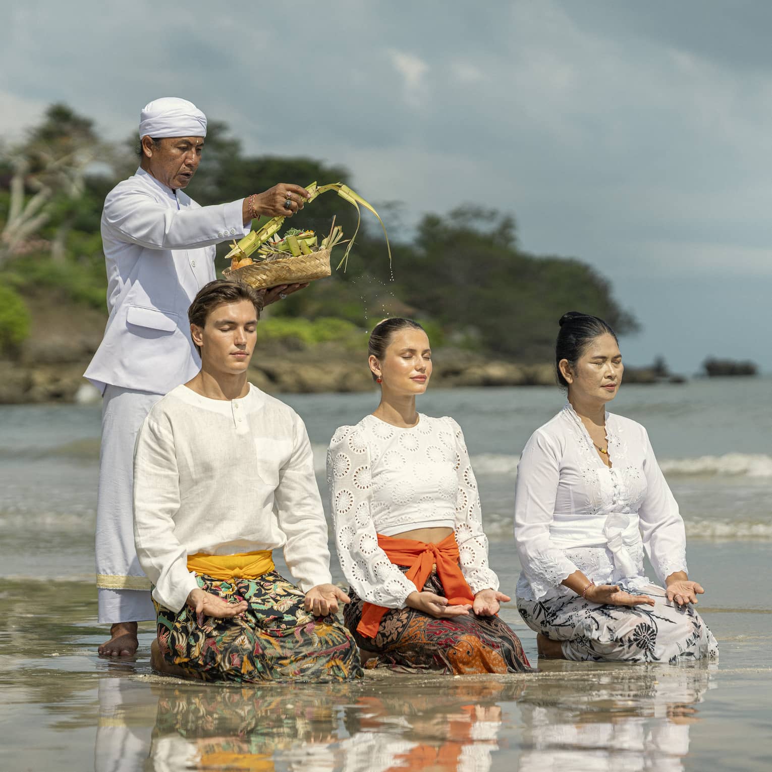 Three adults sit in shallow water with palms up on their knees as another sprinkles water from palm fronds on their heads.