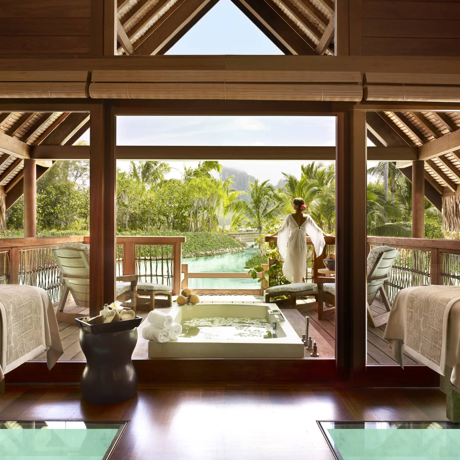Woman wearing flowing white robe stands at edge of spa suite near plunge bath, couples massage beds