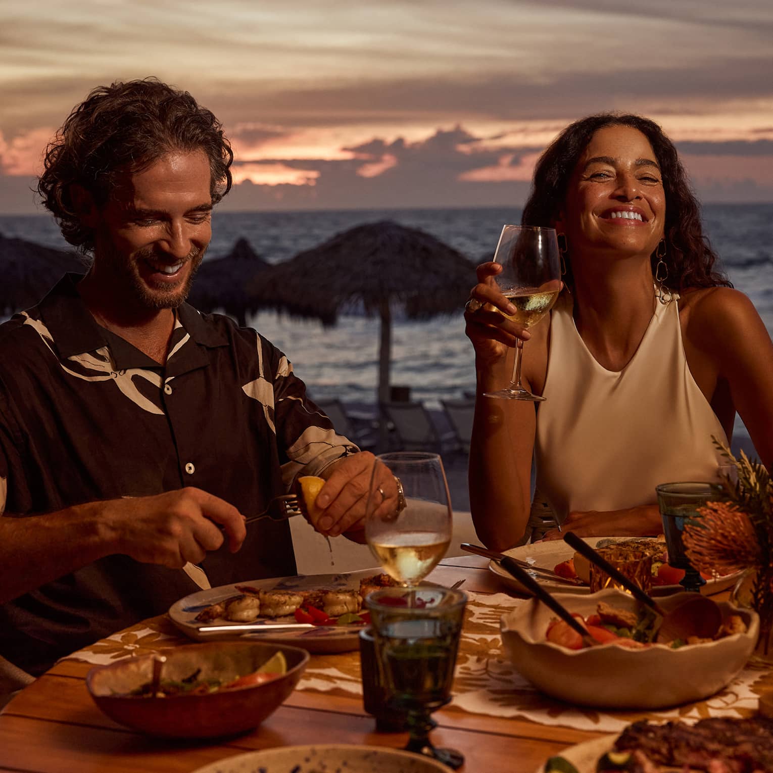 Man and woman laugh while dining at outdoor table filled with food and wine, next to beach and ocean