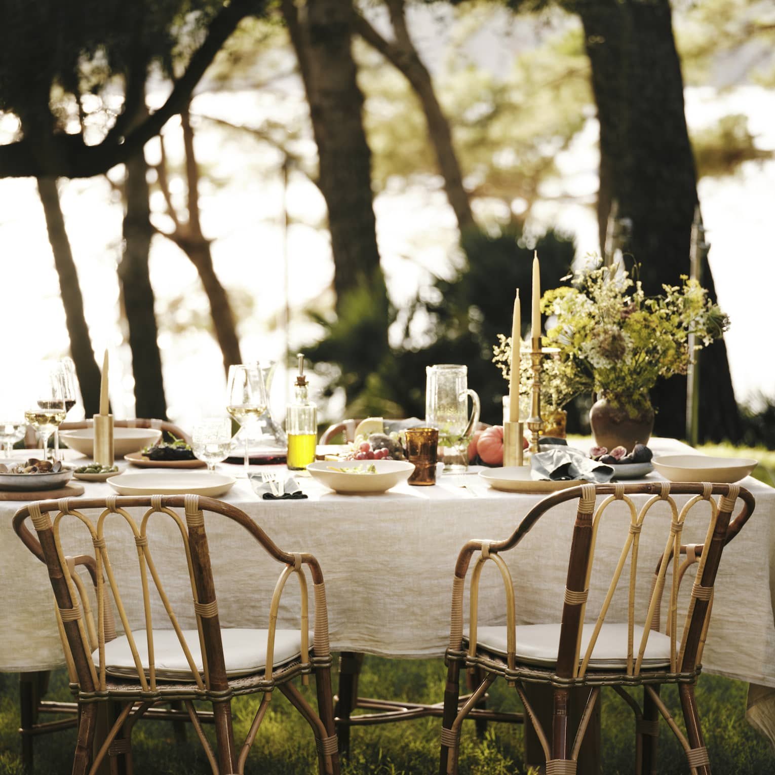 A linen tablecloth adorns an outdoor table set for five with wine, fresh seafood and fruit in an ocean-view forest setting.