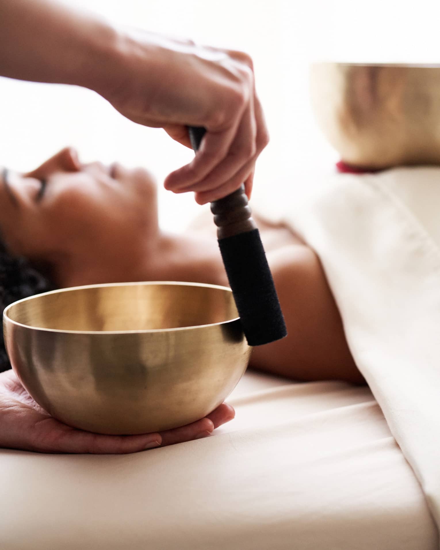 Woman with eyes closed lies on massage table in as vibrational sound therapy is performed with a Tibetan singing bowl