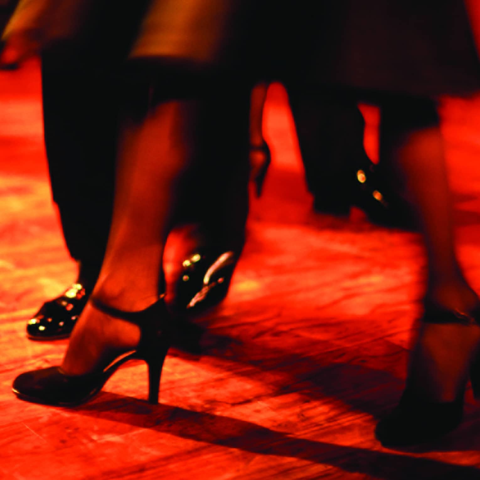 Bathed in a ruby-hued light and amidst other moving legs, a dancer's heel-clad feet step forward in a blur of motion.
