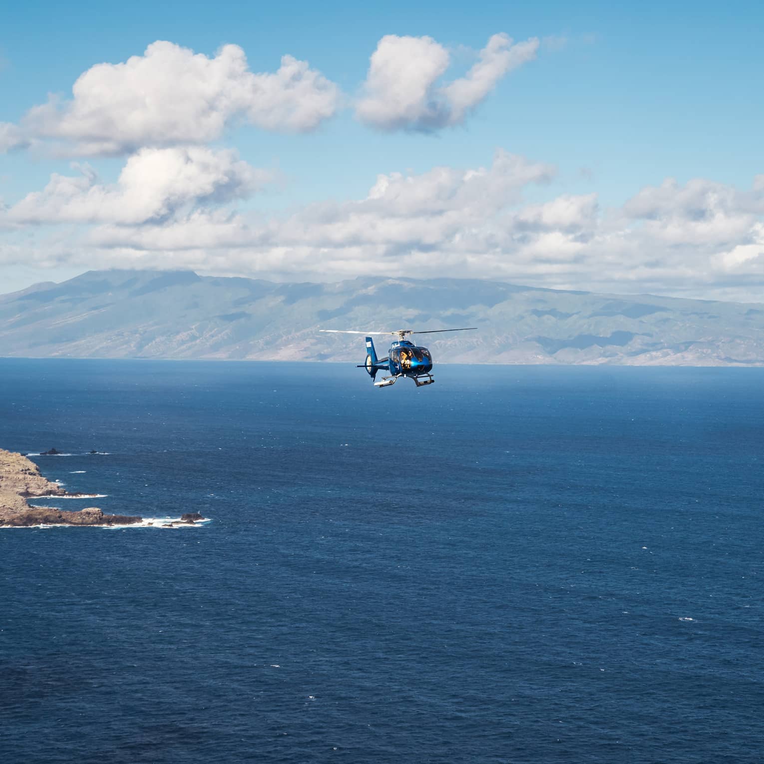 A helicopter flies over open ocean with a mountainous island and blue sky dotted with clouds in the background
