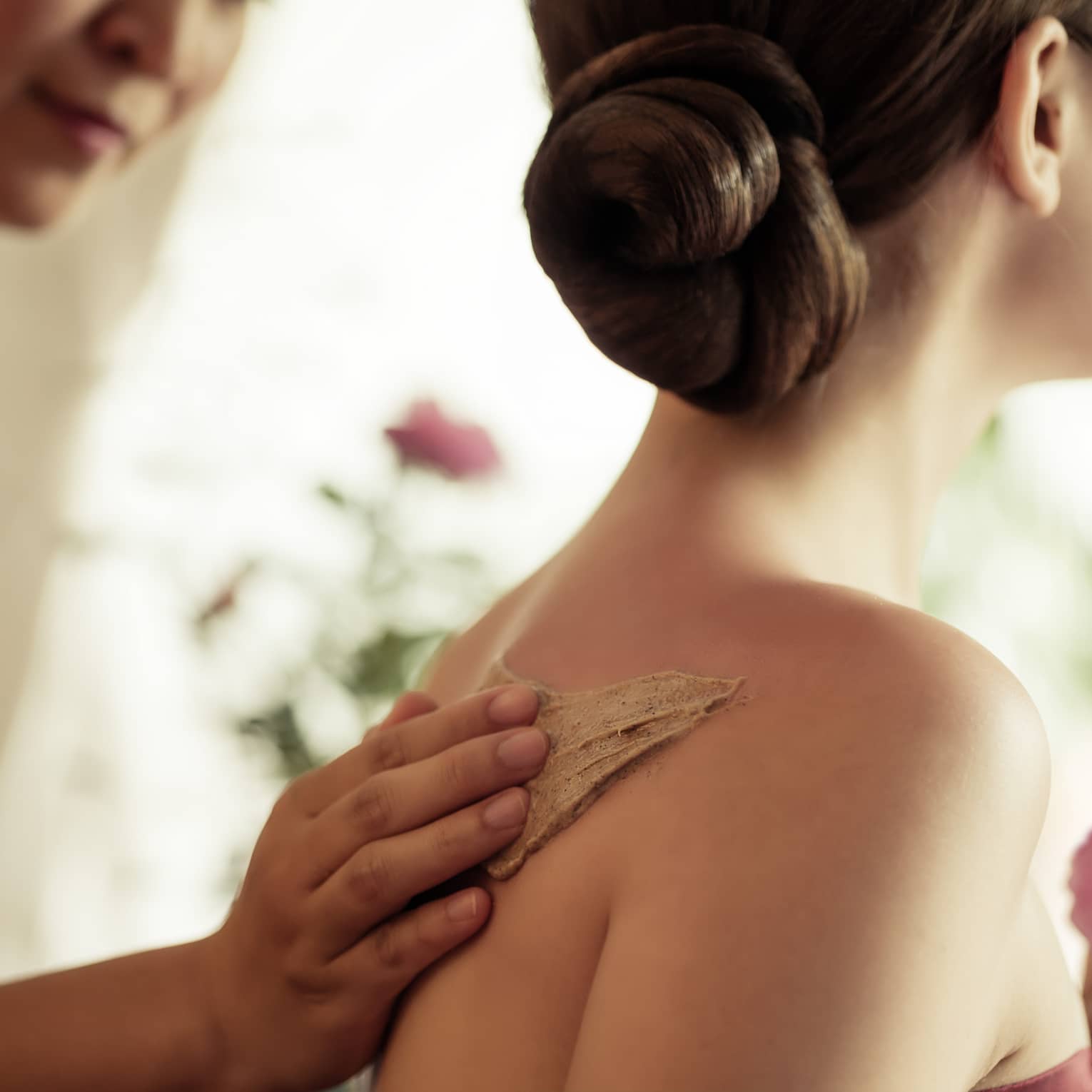 Spa staff rubs thick lotion on woman's bare shoulders as she holds a fresh flower