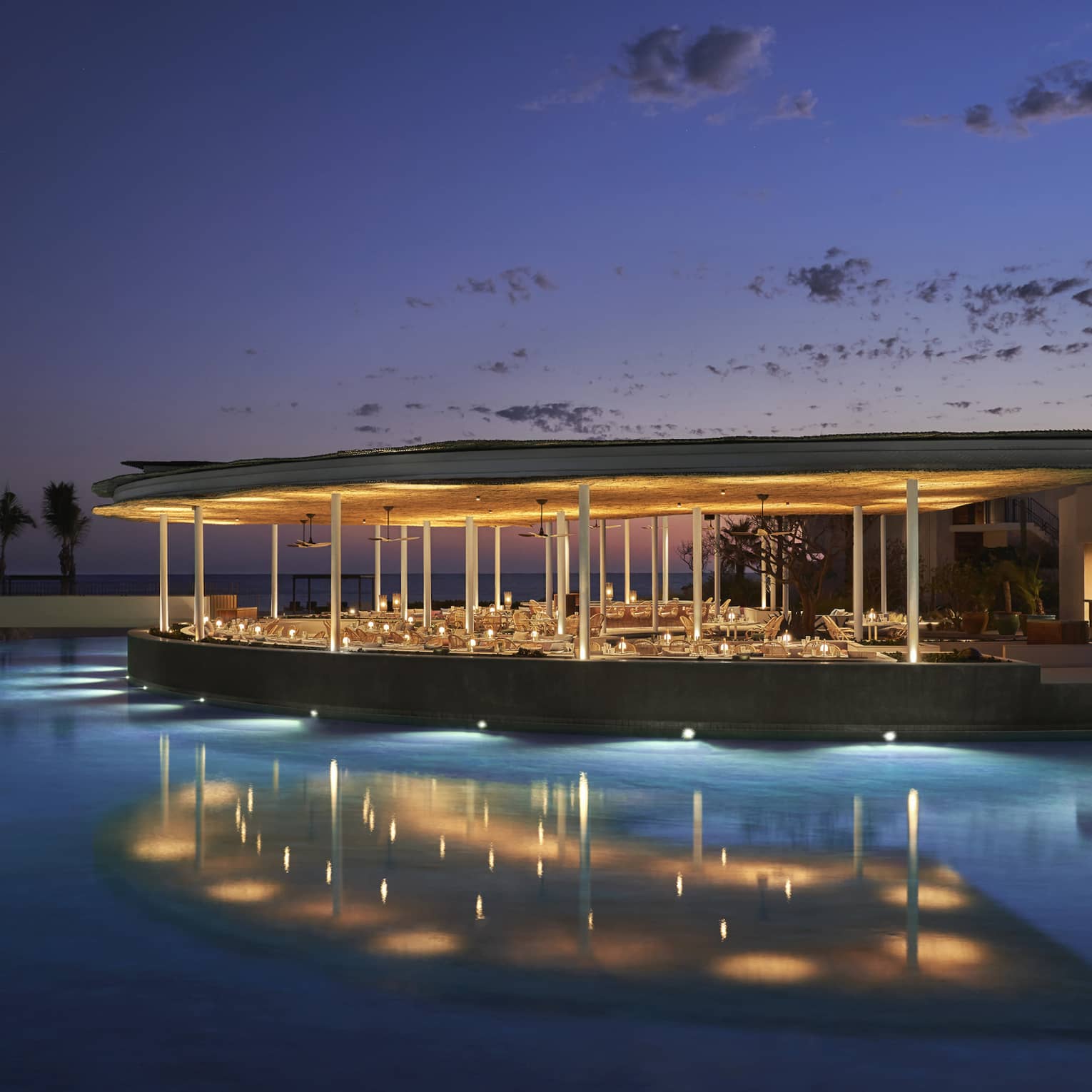 The exterior of an outdoor eating area at night on the water of a shore, it is well lit with lights.