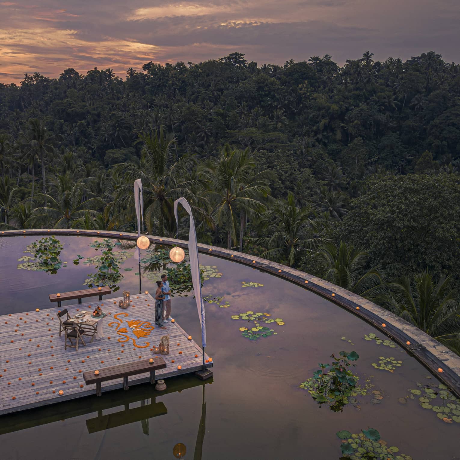 Couple stands on platform over lotus pond at dusk, dining table and hanging lanterns surrounding it, views of lush forest in front of them
