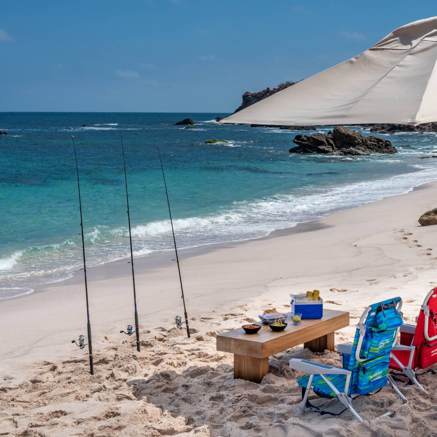 Under an umbrella, drinks, snacks and three colourful beach chairs. Three fishing rods planted in the sand, blue sea ahead.