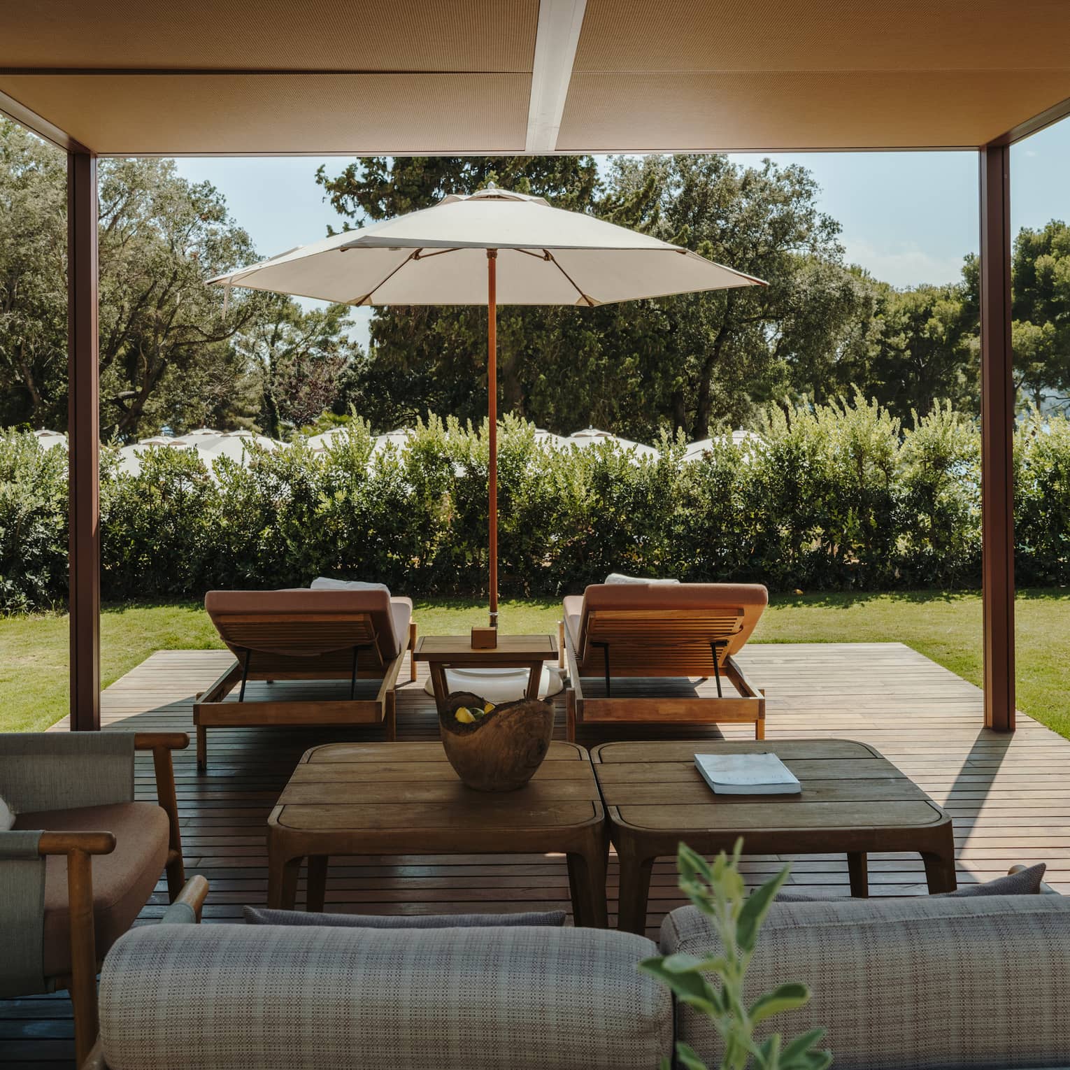 A shaded pool cabana with comfortable seating, a table, lounge chairs, an umbrella and lush green surroundings.
