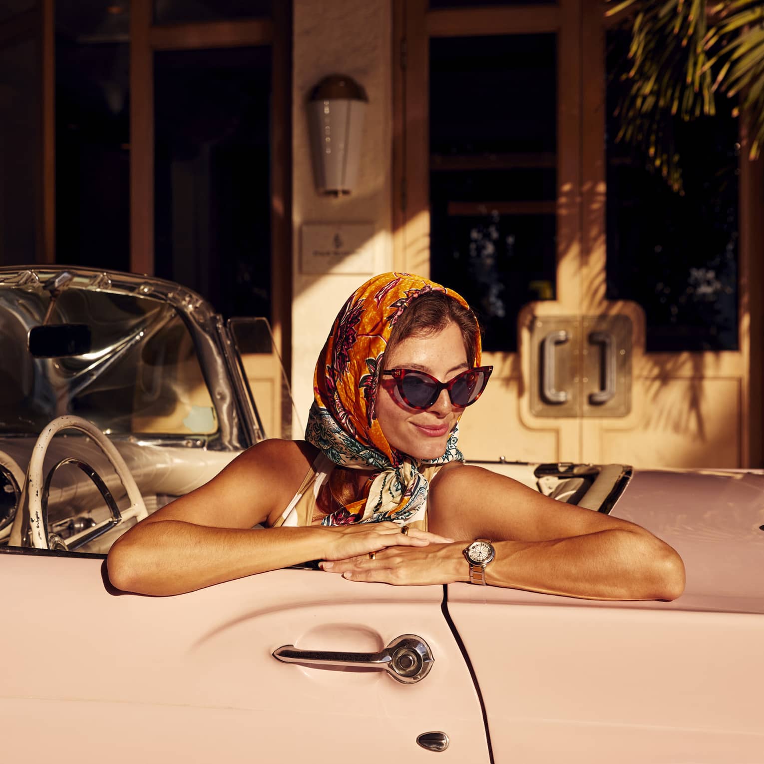Guest wearing a colourful silk head scarf, cat-eye sunglasses, sundress and silver watch sits in a pink convertible