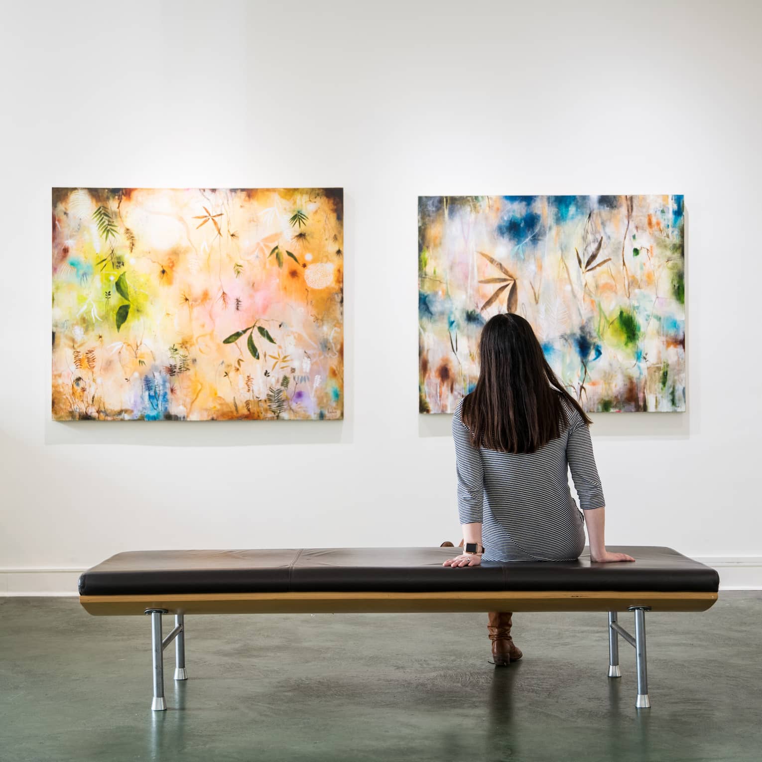 View from behind of an adult seated on a cushioned bench in an art gallery gazing at two brightly coloured modern paintings.