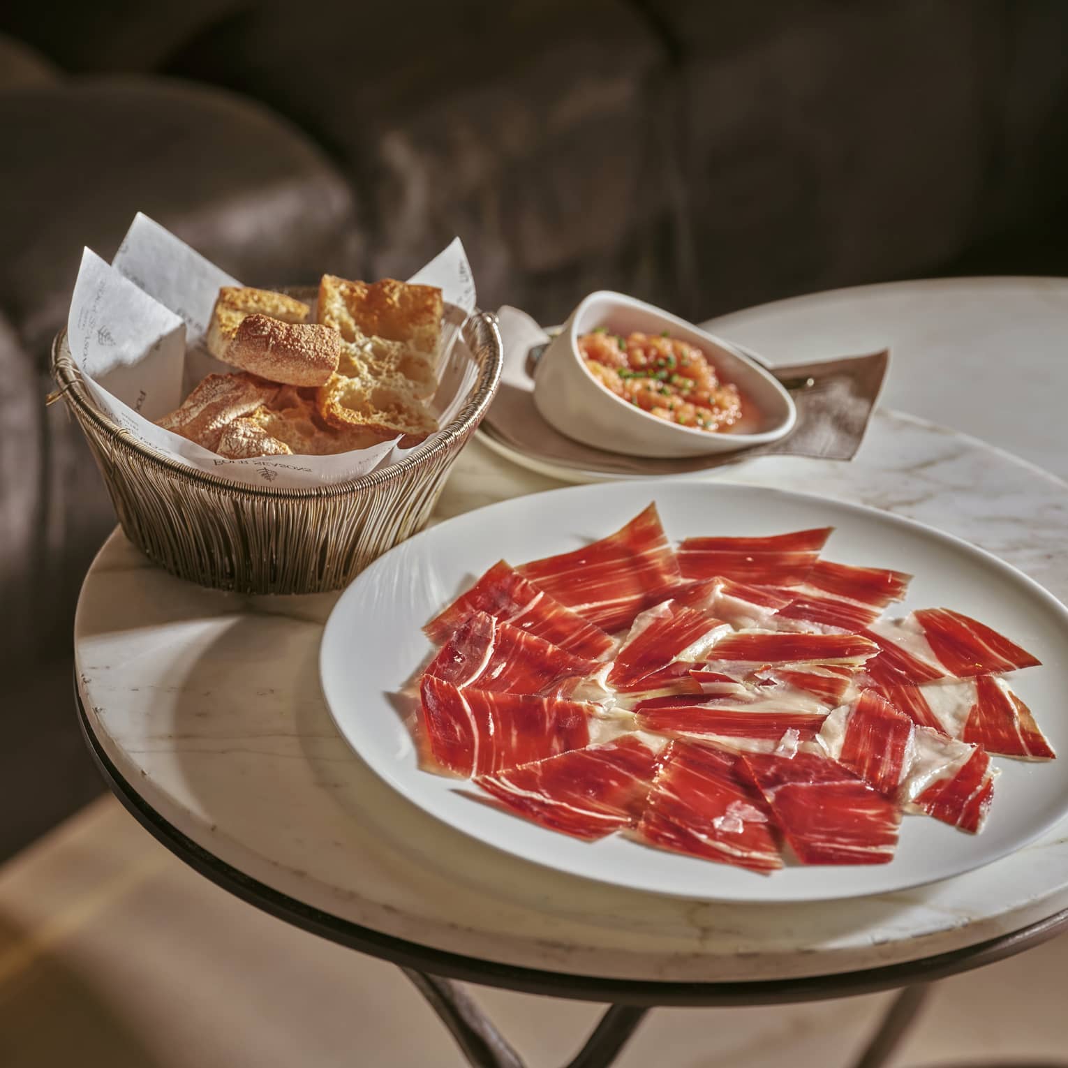 Plate of Iberico ham slices beside bowl of crackers on small marble-top table