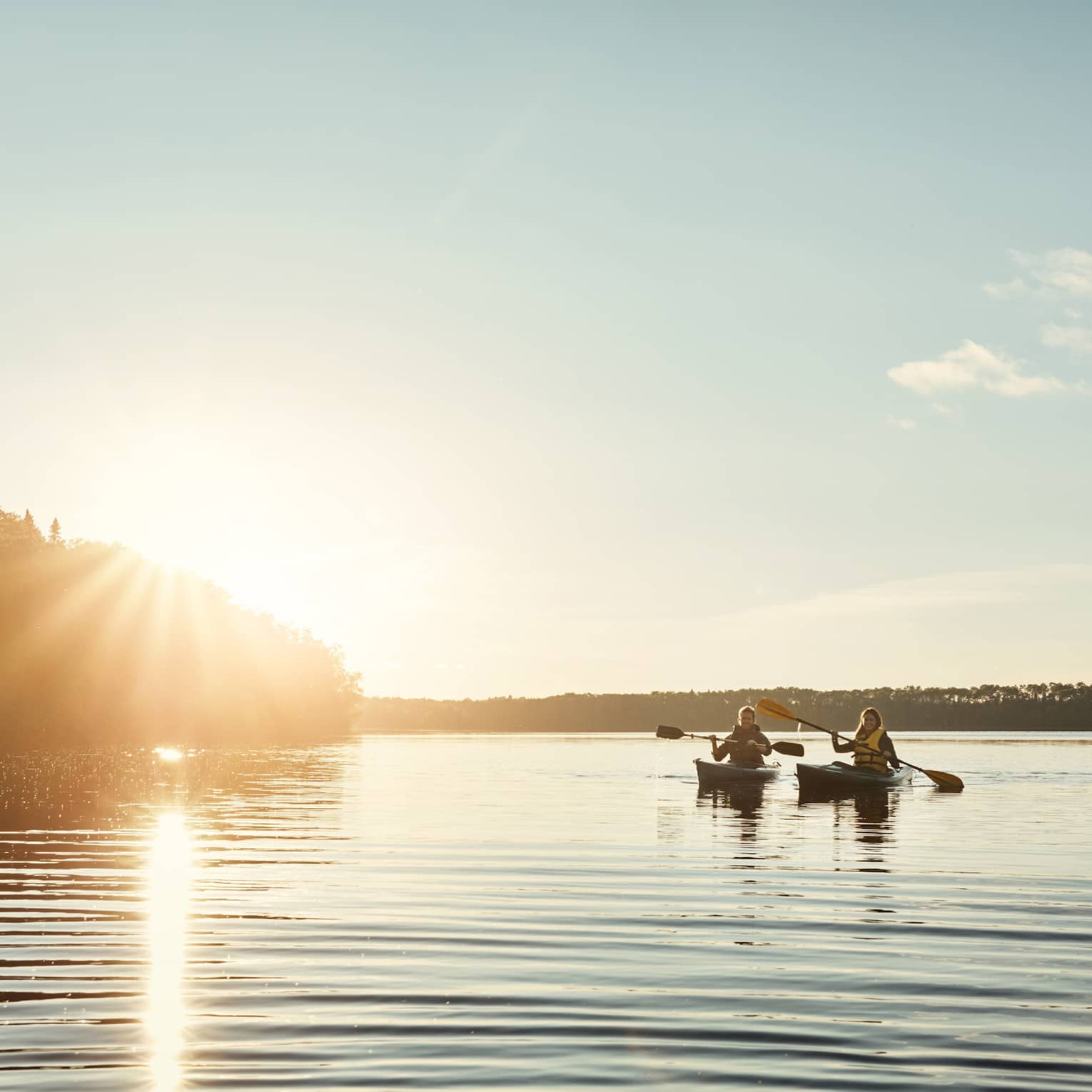 Two people kayaking through a body of water flanked by trees, both silhouetted by the setting sun