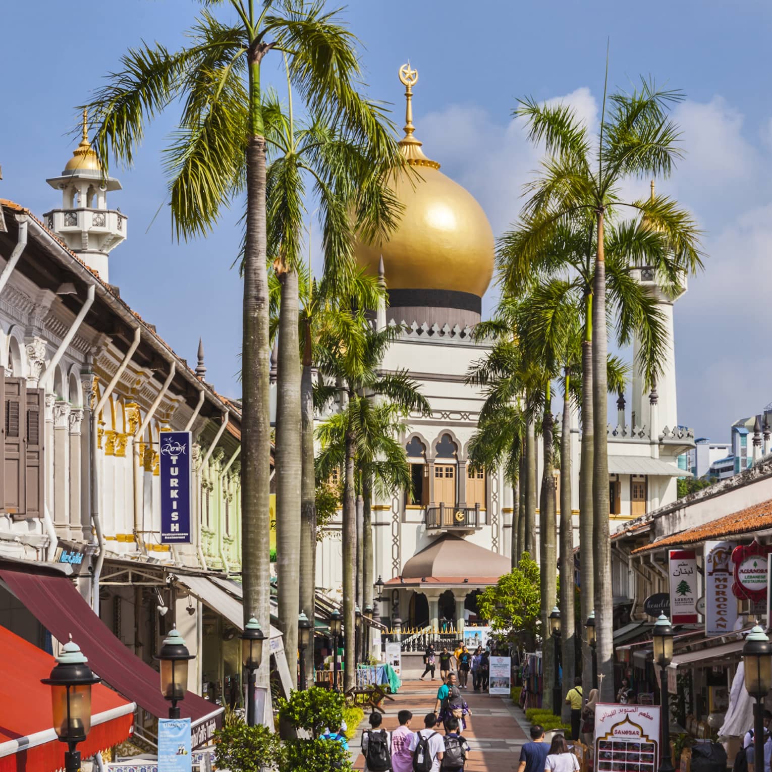 Under a clear blue sky, people mill about a palm tree–lined street of shops that leads to a towering golden-domed mosque