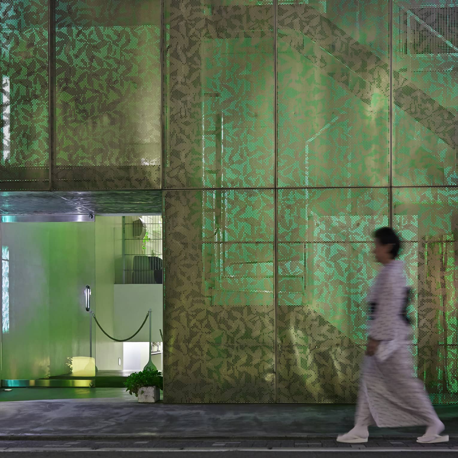 A person heads toward a door to a modern room, passing a multi-paned screened wall with a leaf pattern illuminated in green.