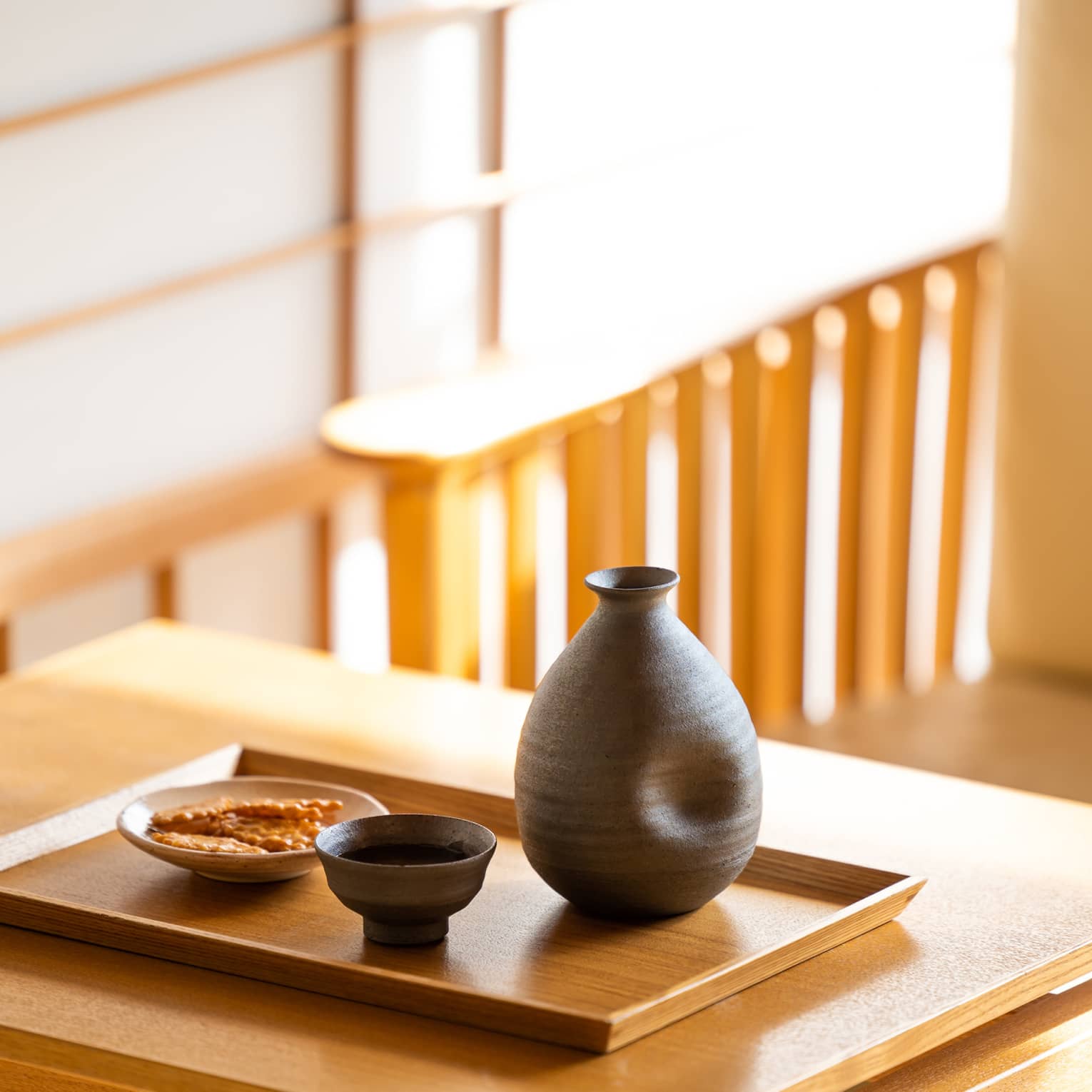 Close-up of a dark ceramic sake carafe with matching cup and a small plate of crackers on a wooden tray and table.