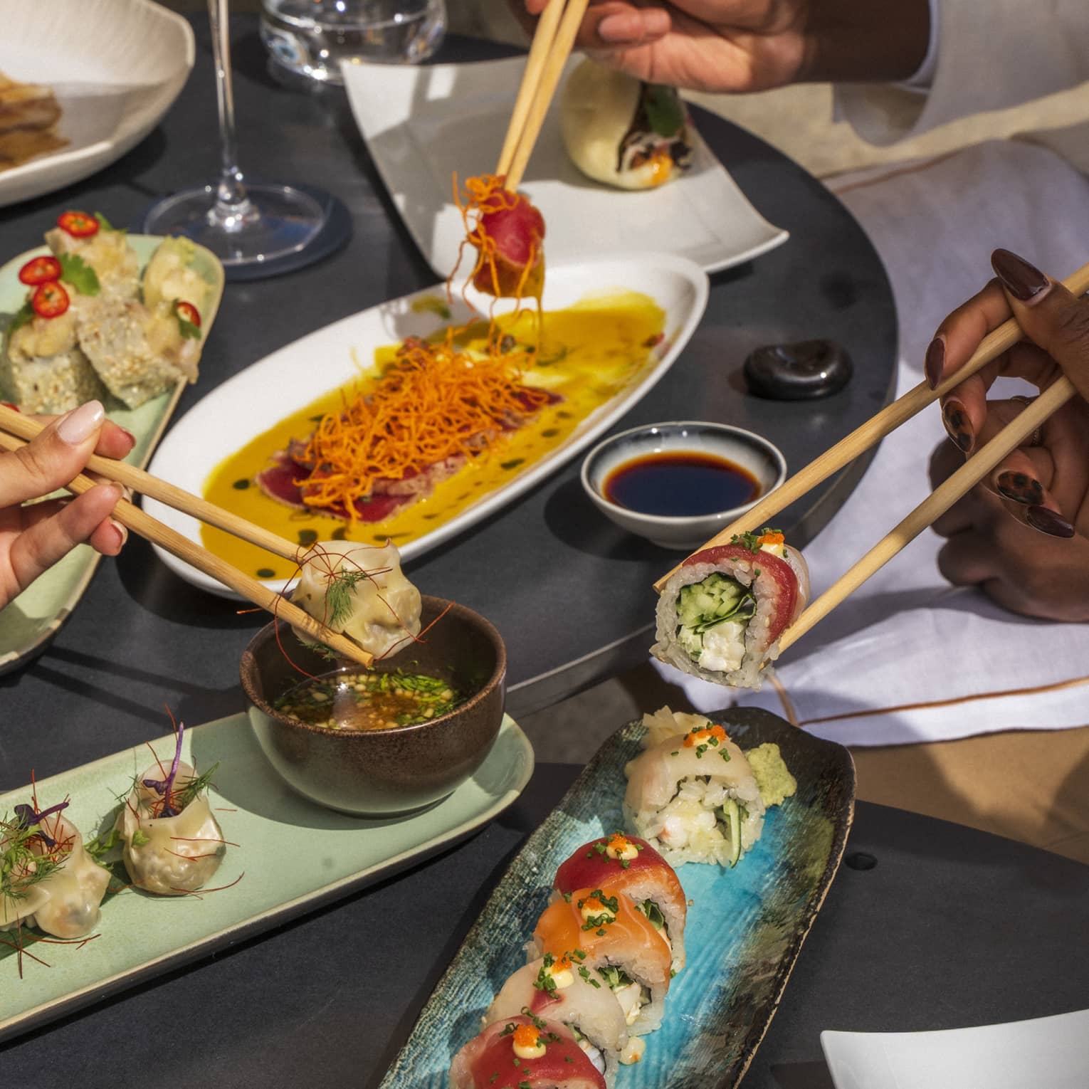 Three hands picking up sushi with chopsticks over plates of dishes