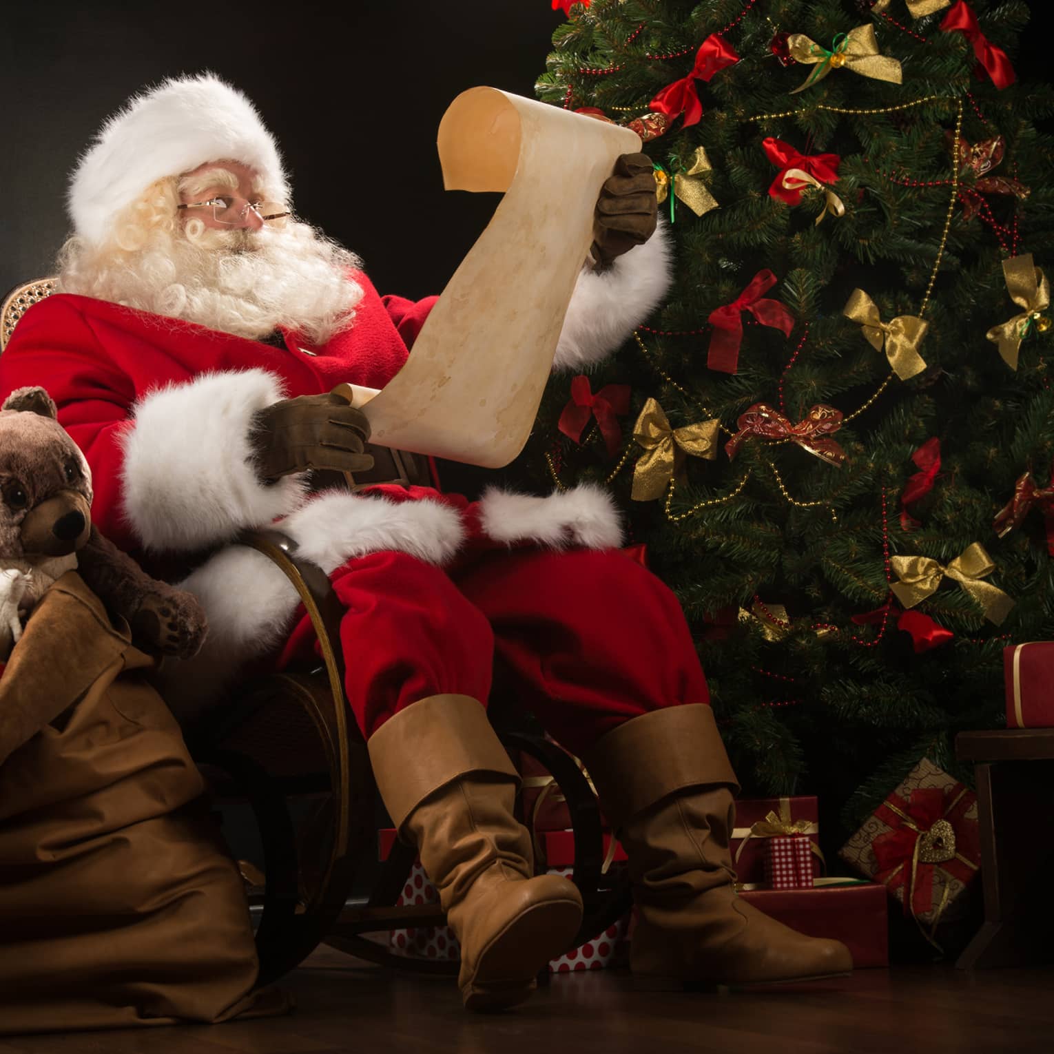 Santa Claus seated in large rocker holding a scroll, flanked by decorated Christmas tree with gifts and overflowing gift bag