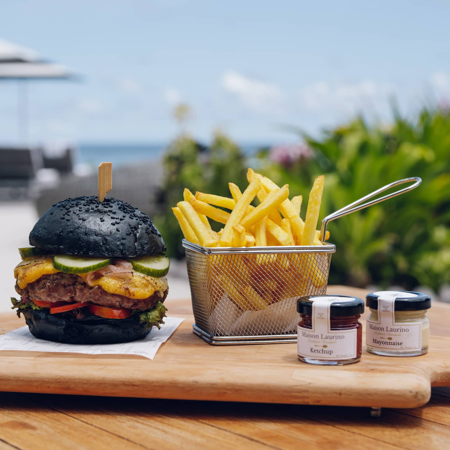 Burger with black bun and fries in metal basket on wooden serving board by the ocean