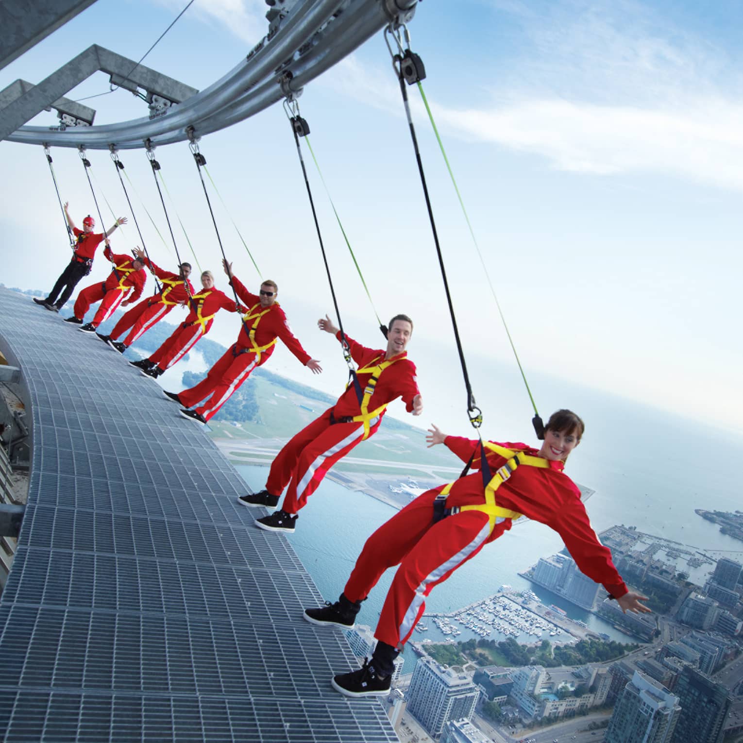 People wearing red jumpsuits, harnesses lean back over edge of CN Tower high above city