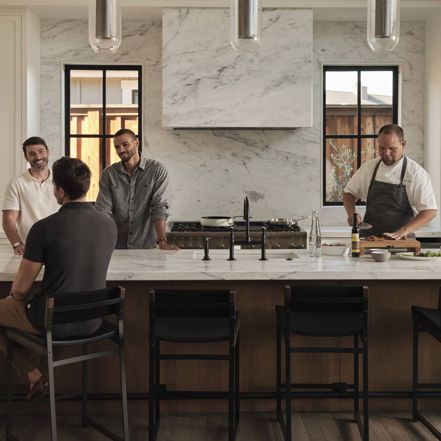A group of guests gather at one end of a long kitchen island while a chef chops ingredients at the other end of the island.