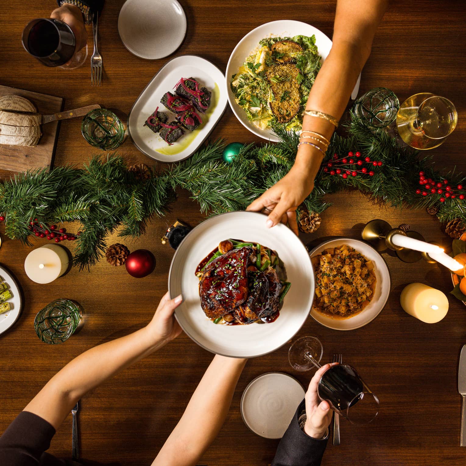 Hands pass a dish of food to another over a table set for a holiday meal with a green garland in the centre