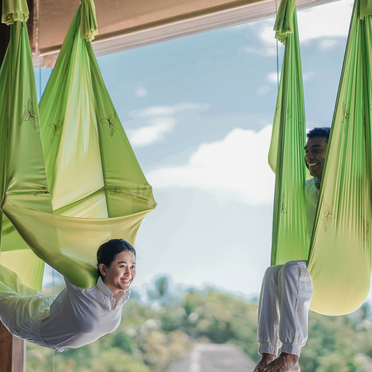 Two people practising antigravity yoga in bright green hammocks, suspended in a wellness resort studio with a view of lush tropical surroundings