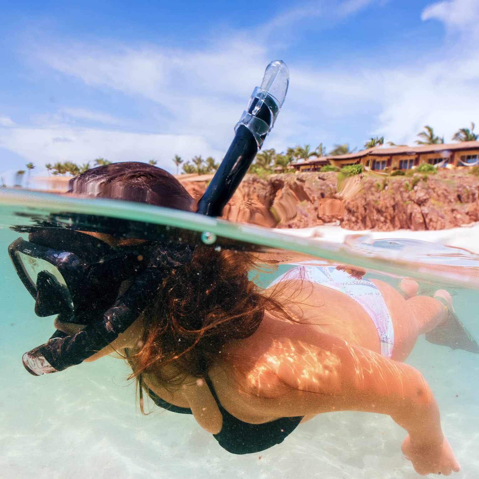 A snorkeller beneath the surface of clear aqua water. Behind, a white sandy beach, swaying palm trees and low-rise buildings.