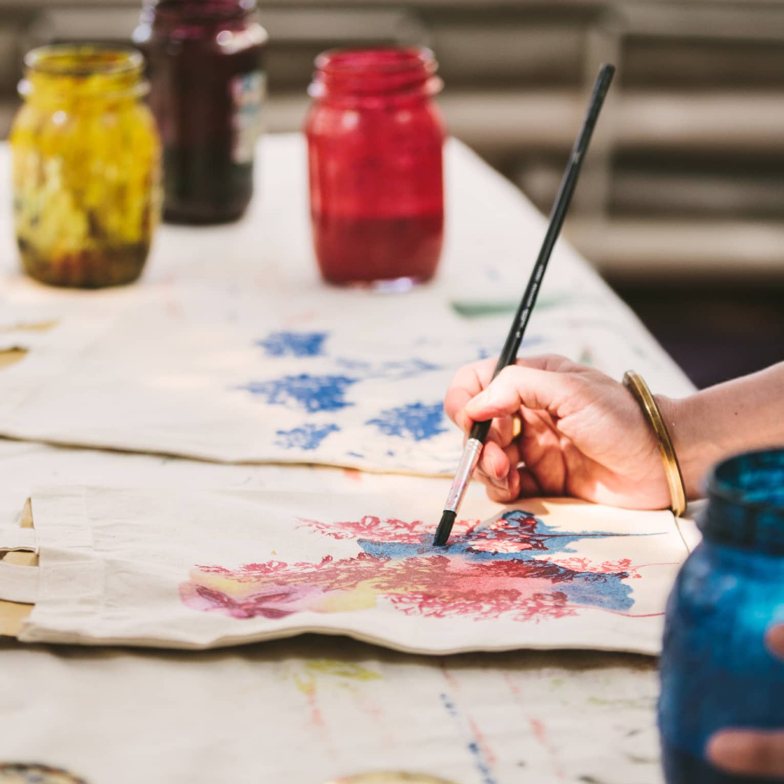 An artist using mason jars filled with red, blue, yellow and black paint to paint images of flowers on canvas tote bags.