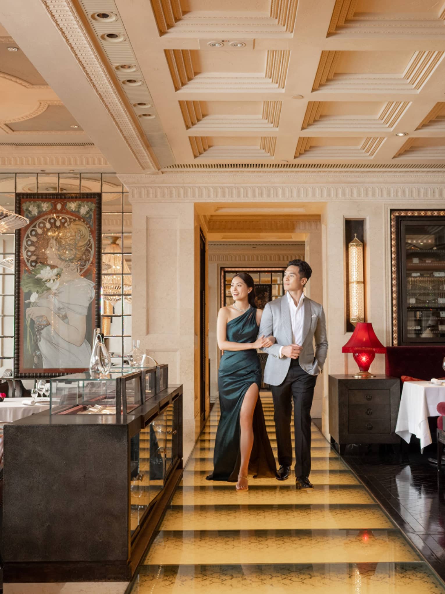 Elegant couple walking arm-in-arm in luxurious restaurant with chandeliers, red velvet seating and ornate decor.