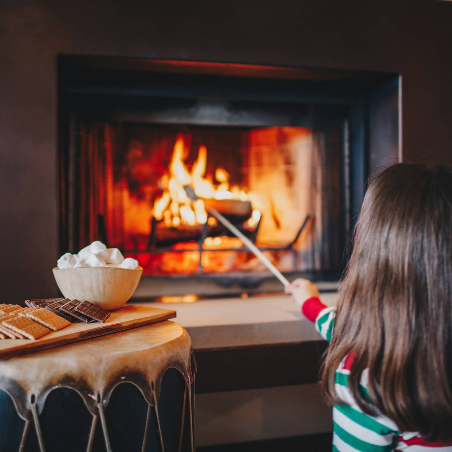 A little girl in red and green pajamas roasts marshmallows in a fireplace