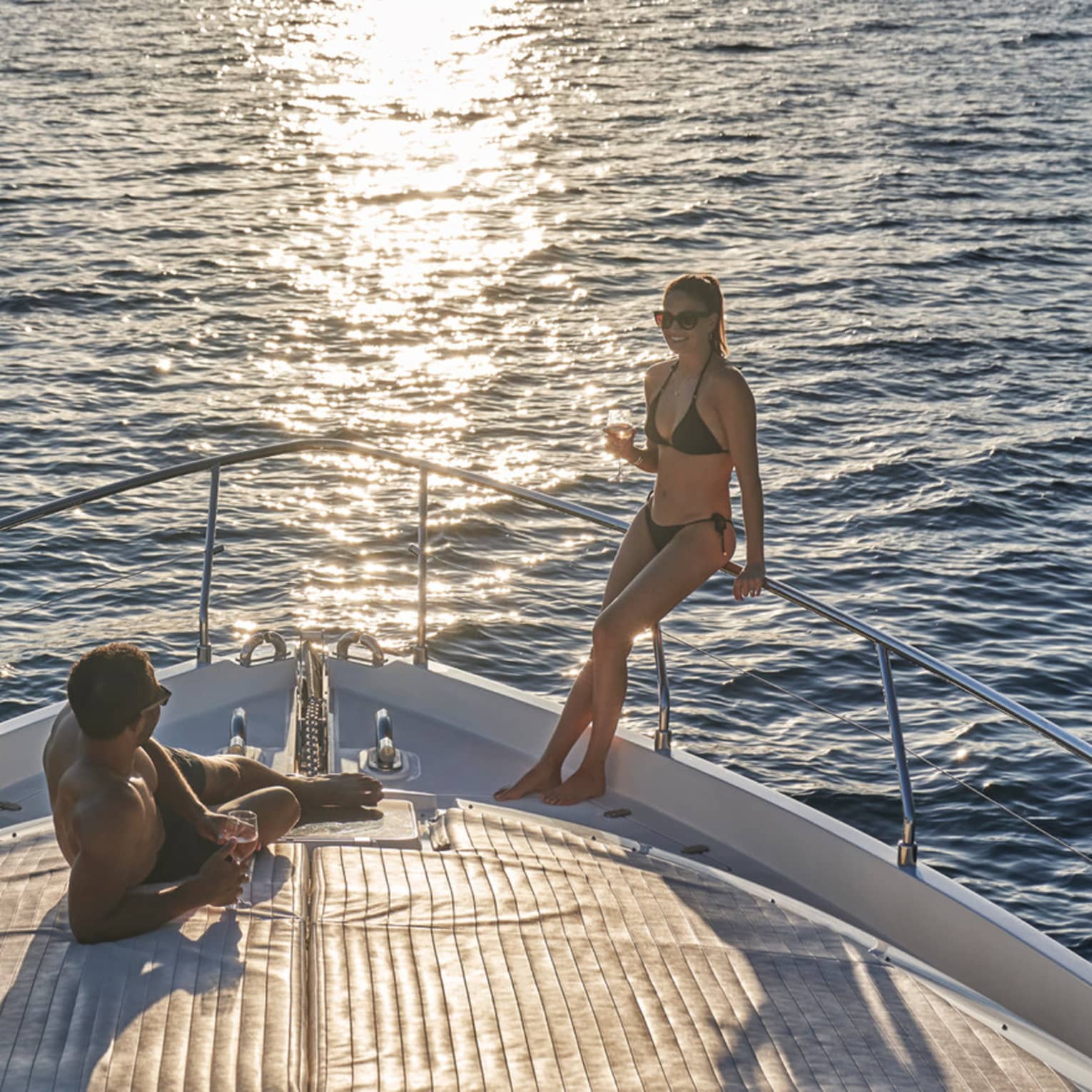 A couple in swimwear relaxing on the bow of a boat, wine glasses in hand and the setting sun glinting off the endless ocean.