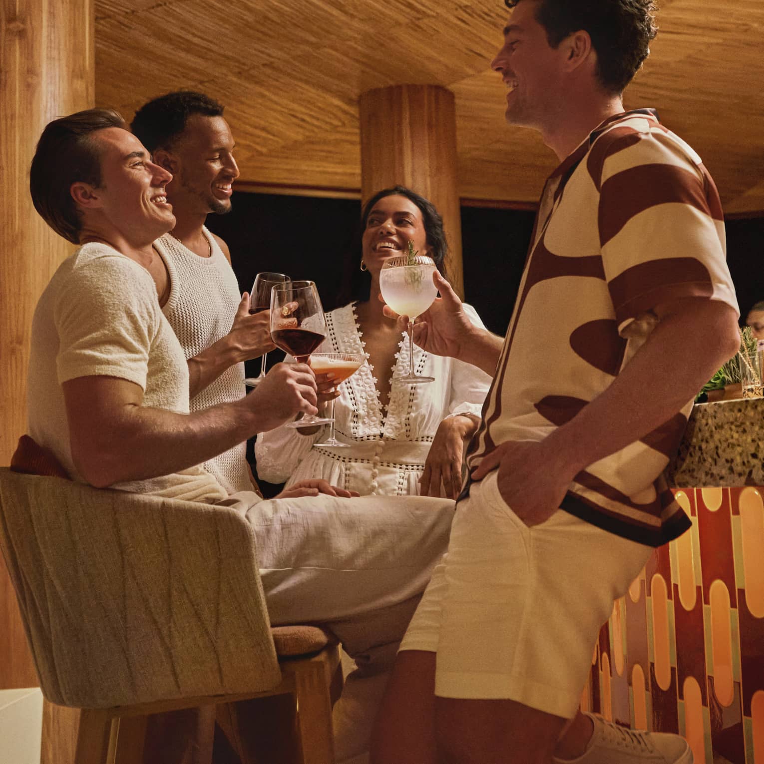 Four guests in stylish summer clothes hold cocktails and socialize next to a terazzo-topped bar in a round wooden pavilion.