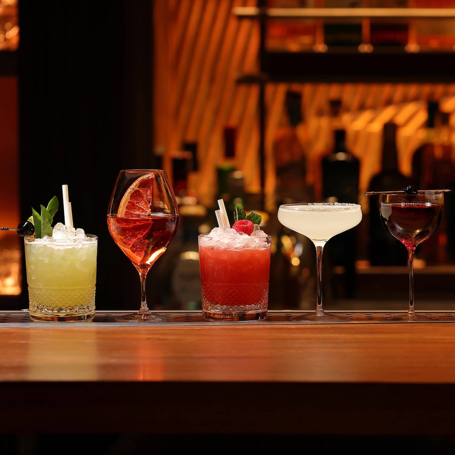 A line of cocktails in various tumblers and stemmed glasses against a backdrop of blurred liquor bottles in reddish lighting.