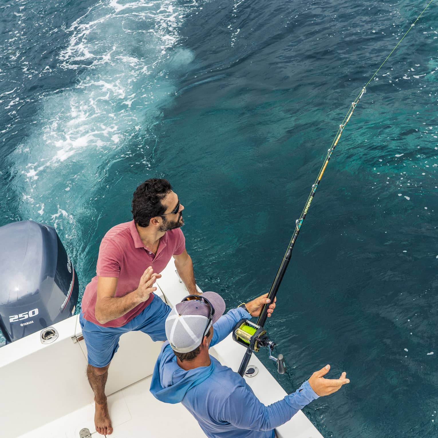 Aerial view of two people fishing off the back corner of a motorboat. One grips a fishing rod while the other looks on.