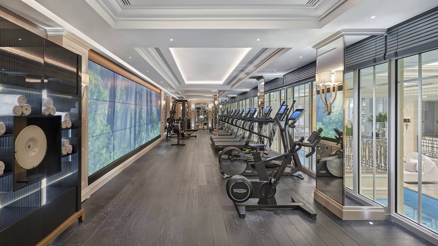 A row of cardio machines in an indoor gym, with windows looking out to indoor pool area
