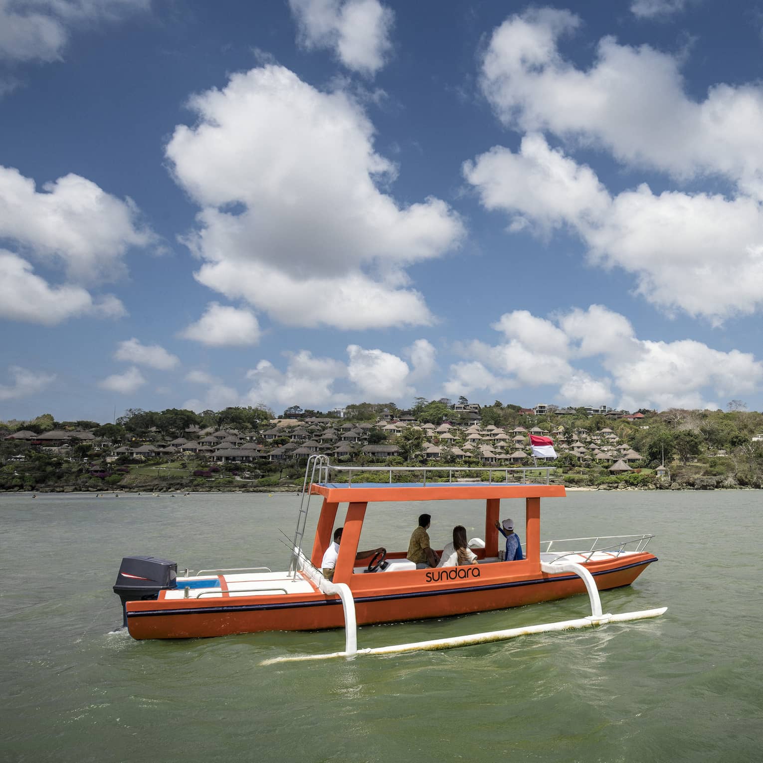 An outrigger canoe glides along calm, green water as four passengers look toward a distant shore dotted with trees and huts.